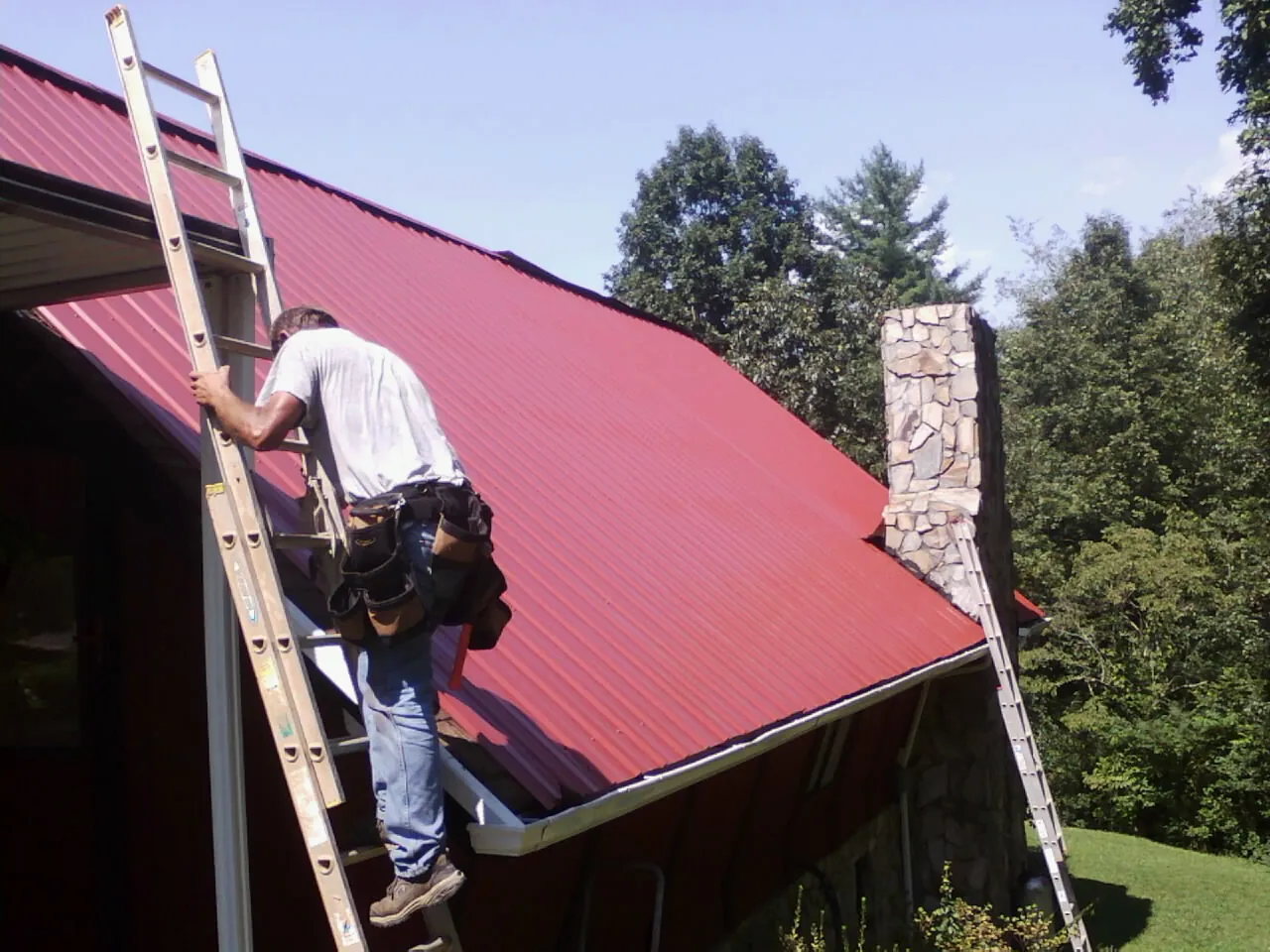 Roofer on a ladder beside a red metal roof and stone chimney — Soffit & Fascia Repair by Roofing Brothers in Ingleside